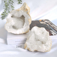 Two large crystal geodes on a white surface with a book and plant in the background.