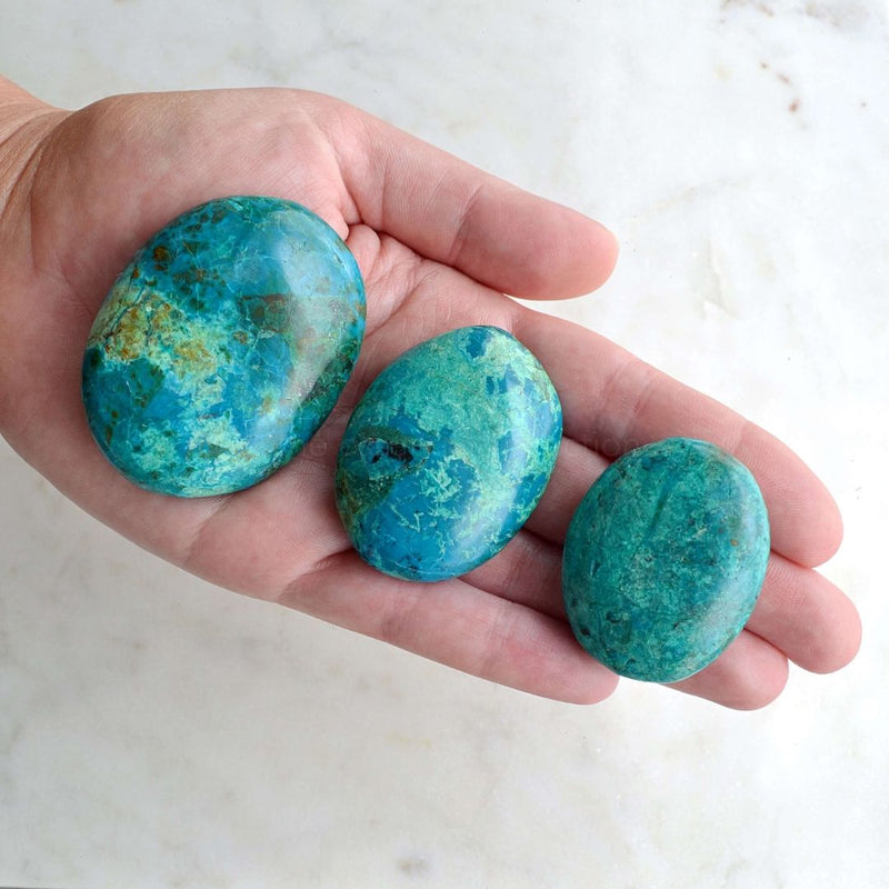 White marble background with hand holding 3 different sizes of Chrysocolla Palm Stones which each contain a mix of colours, greens, blues, blacks. 
